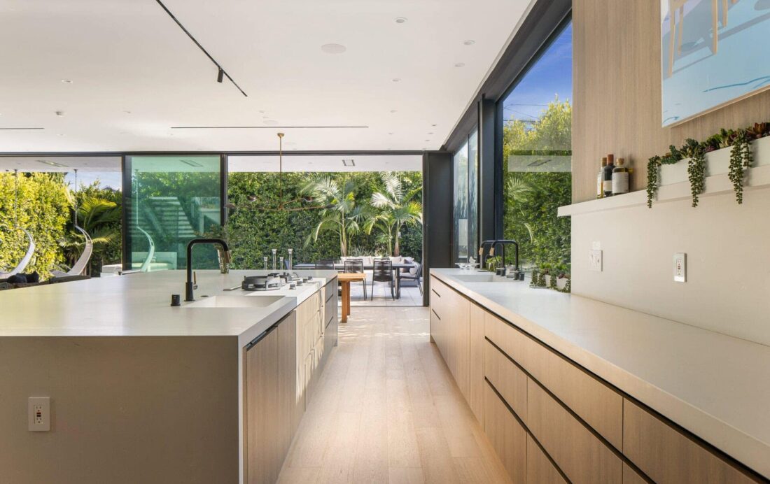 Contemporary kitchen with minimalist design, white surfaces, natural wood cabinetry, expansive glass sliding doors showing lush greenery outside.