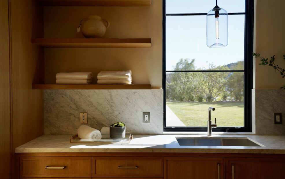 Sunlit laundry room with oak cabinetry, marble countertops, and a large window overlooking a landscaped yard