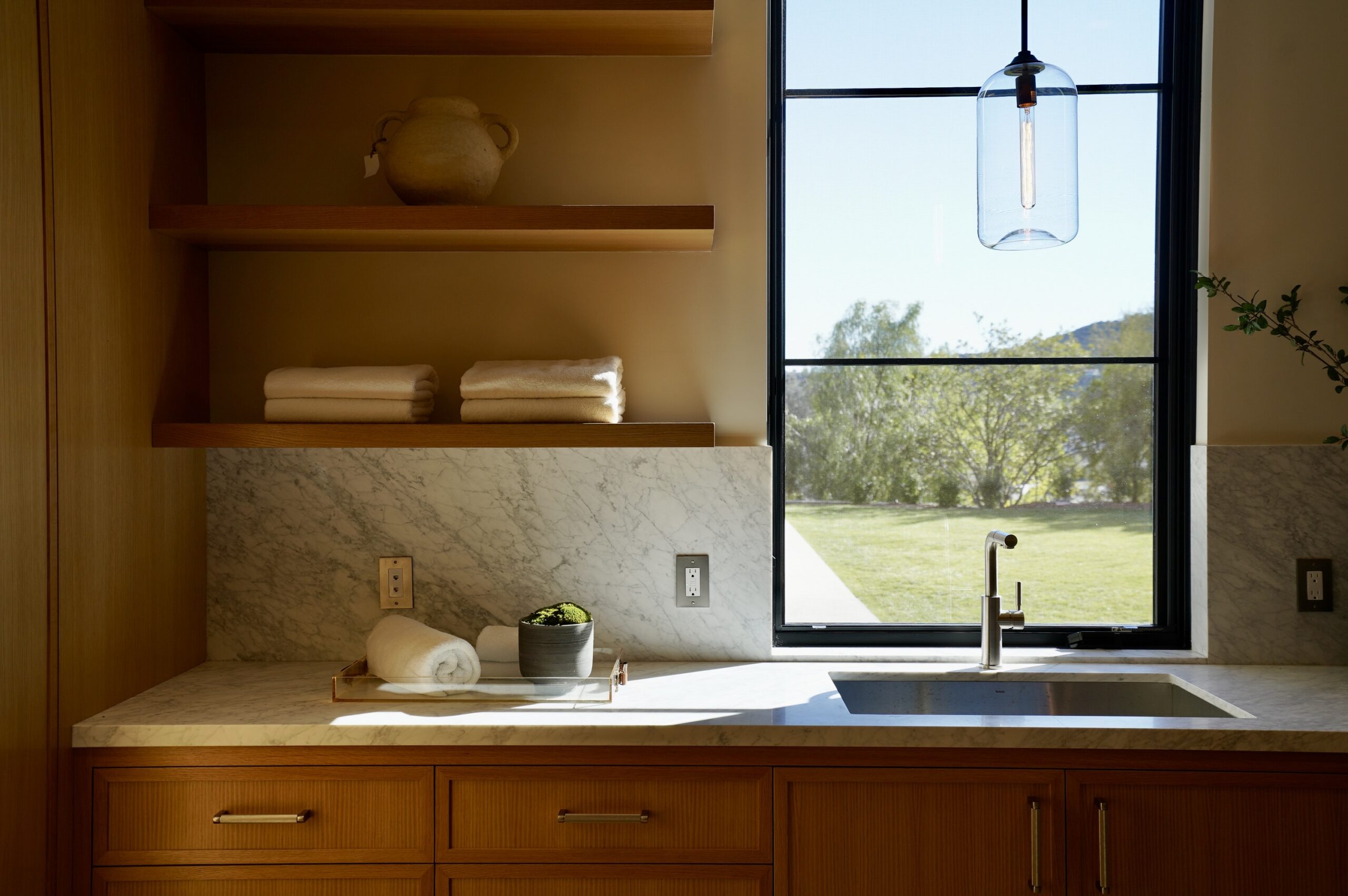 Sunlit laundry room with oak cabinetry, marble countertops, and a large window overlooking a landscaped yard